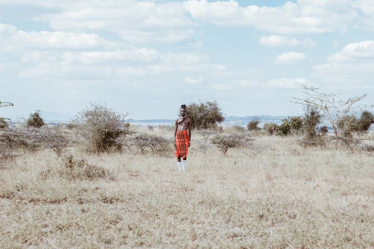 A person in traditional Kenyan attire stands in a vast grass field under a cloudy sky.