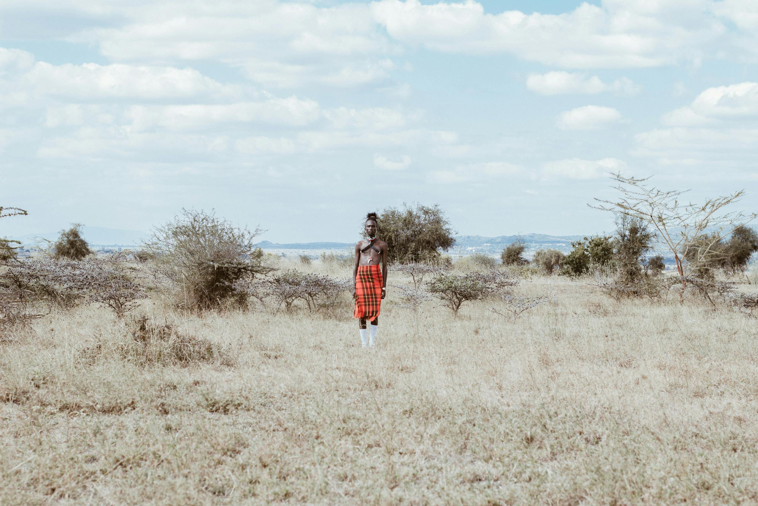 A person in traditional Kenyan attire stands in a vast grass field under a cloudy sky.